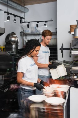 young interracial volunteers in t-shirts with lettering holding plastic containers in kitchen