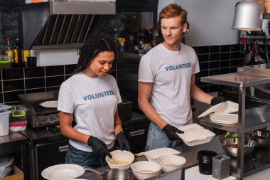 young multiethnic volunteers in t-shirts with lettering holding plastic containers in kitchen