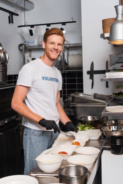 happy and redhead volunteer in latex gloves holding tweezers near plastic containers in kitchen 