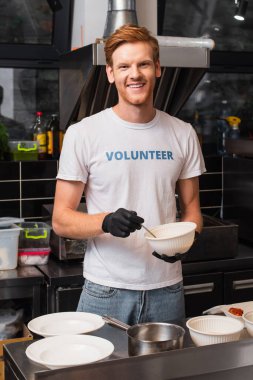happy and redhead volunteer in latex gloves holding plastic bowl in kitchen 