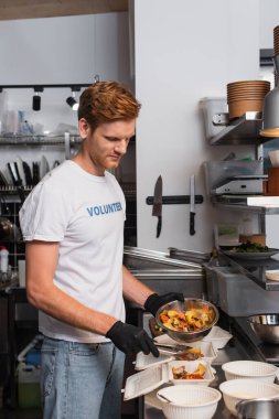 redhead social worker in latex gloves holding bowl with prepared food near plastic containers