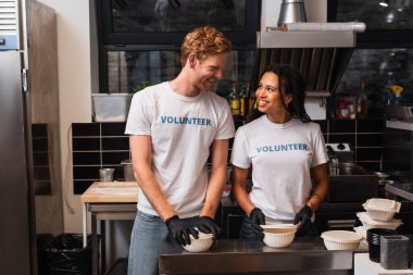 happy multiethnic volunteers in t-shirts preparing plastic containers with food in kitchen 