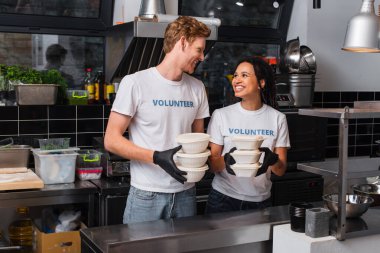 happy multiethnic volunteers in t-shirts holding plastic containers with food in kitchen  clipart