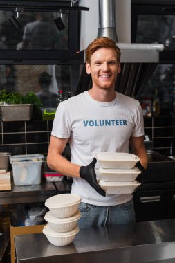 redhead man in t-shirt with volunteer lettering smiling and holding plastic containers in kitchen 