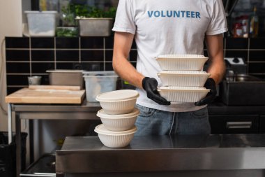 cropped view of man in t-shirt with volunteer lettering holding plastic containers in kitchen 