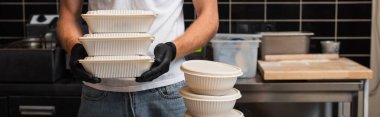 cropped view of man in t-shirt with volunteer lettering holding plastic containers in kitchen, banner
