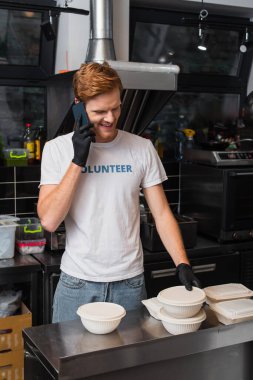 redhead man in t-shirt with volunteer lettering smiling and holding plastic bowl and talking on smartphone in kitchen 