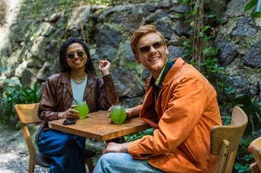 happy multiethnic friends in sunglasses and jackets holding glasses with green alcohol cocktail in outdoor terrace