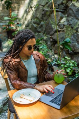 young african american woman in sunglasses using laptop near prepared lunch and glass with drink in outdoor cafe terrace