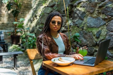 young african american freelancer in sunglasses using laptop near prepared lunch and glass with drink in outdoor cafe terrace