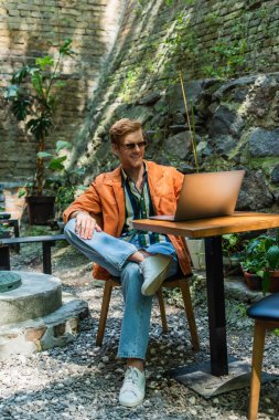happy redhead man in sunglasses working remotely on laptop while sitting in outdoor terrace