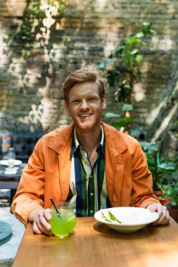 happy redhead man near prepared lunch and glass with cocktail in outdoor cafe terrace