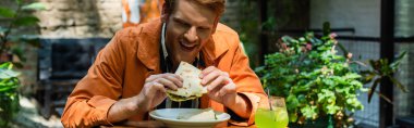 cheerful redhead man eating quesadilla near glass with cocktail in outdoor terrace, banner
