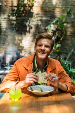 cheerful redhead man holding quesadilla near glass with cocktail in outdoor cafe terrace