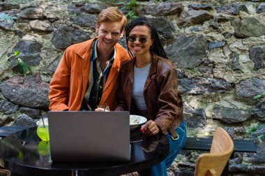 cheerful interracial friends watching movie while having lunch in cafe on terrace 