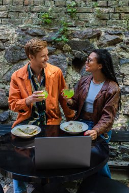 cheerful interracial friends holding glasses with cocktail while having lunch near laptop in cafe on terrace 