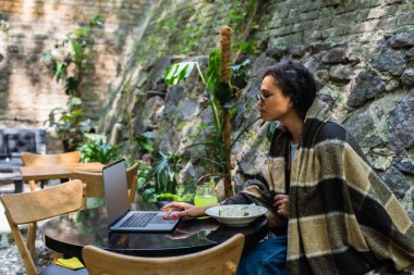 african american freelancer covered in blanket using laptop and having lunch in cafe on terrace