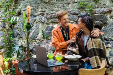 happy redhead man looking at african american woman covered in blanket while having lunch in cafe near laptop