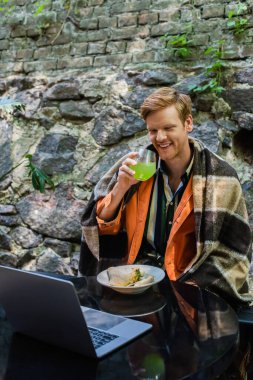 cheerful redhead man covered in blanket drinking cocktail during lunch and looking at laptop