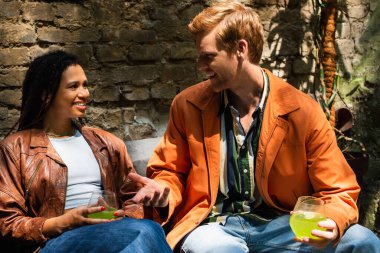 happy multiethnic friends holding glasses with cocktail while talking outside