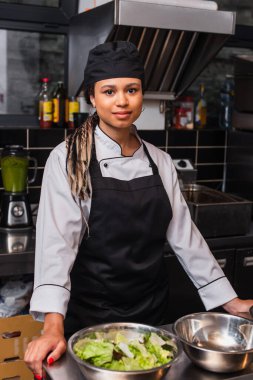 cheerful african american chef in apron standing near bowls and lettuce in kitchen 