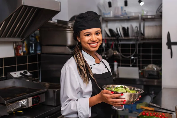 happy and young african american chef holding bowl with salad in professional kitchen