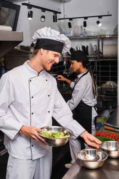happy chef holding bowl with salad near vegetables and african american sous chef in kitchen 