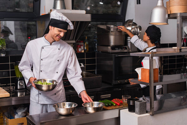 cheerful chef holding bowl with salad near vegetables and african american sous chef in kitchen 