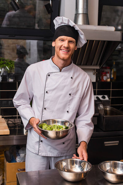 cheerful chef in hat and uniform holding bowl with lettuce  in kitchen 