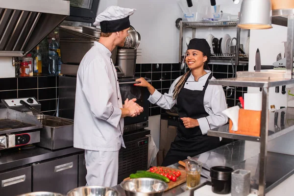 chef in hat and uniform looking at cheerful african american colleague near convection oven