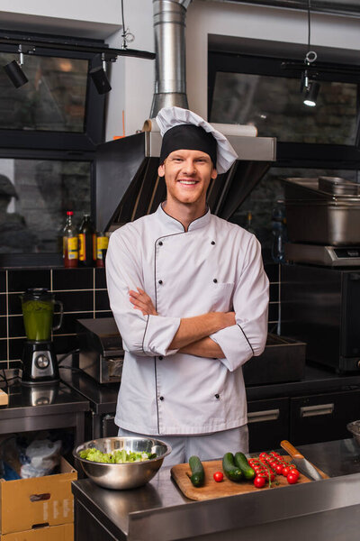happy young chef in uniform standing with crossed arms near vegetables on cutting board in kitchen 