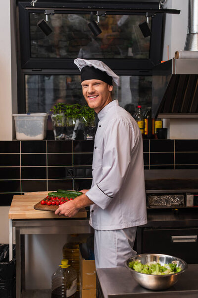 happy young chef in uniform holding cutting board with vegetables in kitchen 