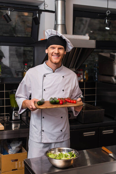 cheerful young chef in uniform holding cutting board with vegetables in kitchen 