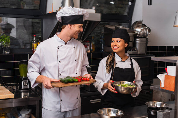 happy chef and cheerful african american sous chef standing with ingredients in kitchen 