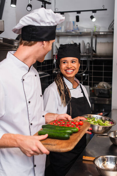 chef and cheerful african american sous chef standing with ingredients in kitchen 