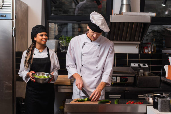 african american woman in apron holding bowl with lettuce and looking at chef preparing meal in kitchen 