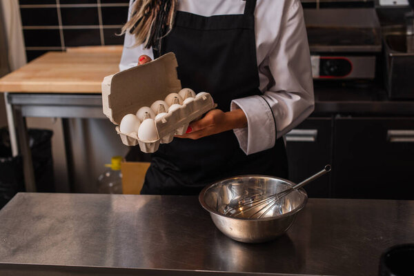 cropped view of african american woman holding chicken eggs in carton near bowl and whisk 