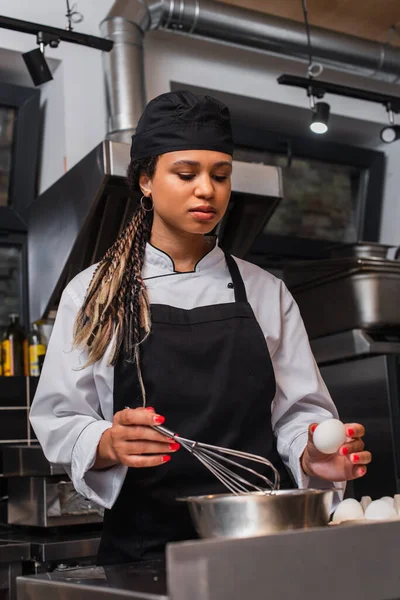 young african american chef in apron holding egg near bowl and whisk in kitchen