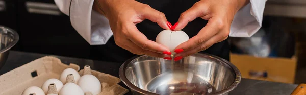 cropped view of african american chef holding raw egg above bowl while cooking in kitchen, banner