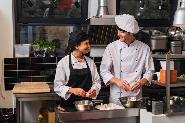cheerful multiethnic chefs smiling while holding raw eggs and cooking in kitchen 