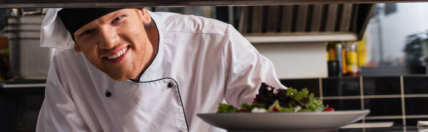 happy chef holding plate with freshly cooked salad in professional kitchen, banner