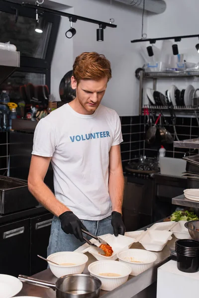redhead volunteer in t-shirt with lettering putting prepared meat in plastic container 