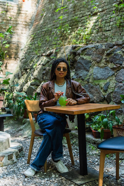young african american woman in sunglasses holding glass with green alcohol cocktail in outdoor terrace 