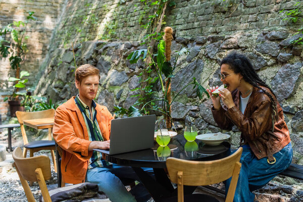 african american woman eating quesadilla near freelancer friend using laptop in outdoor terrace