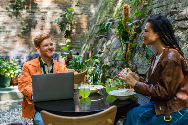african american woman holding quesadilla near happy freelancer friend using laptop in outdoor terrace