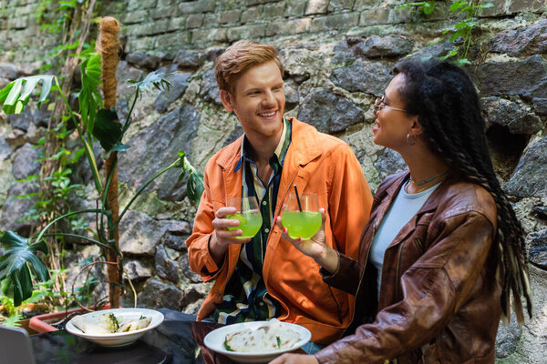 cheerful multiethnic friends holding glasses with cocktail while having lunch near laptop in cafe on terrace 