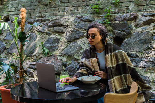 african american freelancer in sunglasses covered in blanket using laptop and having lunch in cafe on terrace