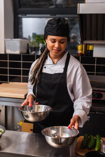 african american chef in apron holding bowl near cucumbers in professional kitchen