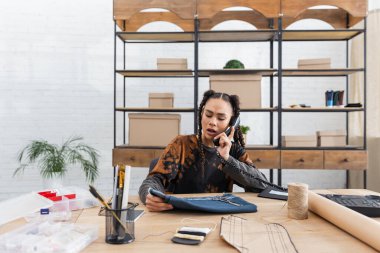 Young african american craftswoman talking on telephone and holding cloth in workshop 