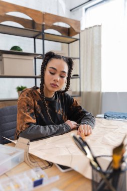 African american designer looking at sewing pattern near blurred equipment in workshop 